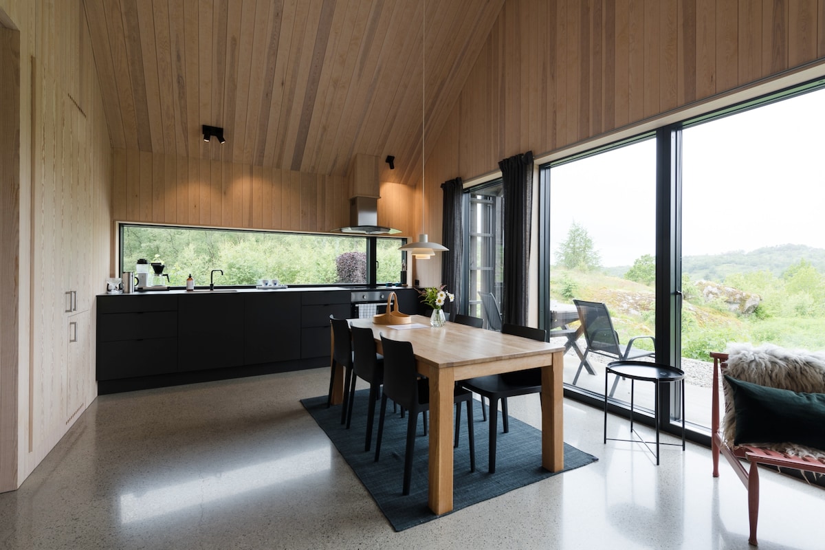 A modern kitchen and dining area is featured, showcasing a wooden dining table surrounded by black chairs. Large windows provide natural light and views of the outdoor landscape, while sleek cabinetry complements the minimalist design of the space.