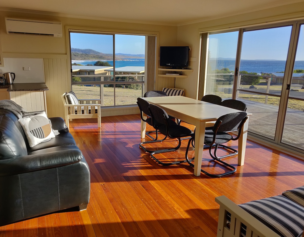 The living area features a spacious table surrounded by six chairs, with a view of the ocean through large glass doors. A dark leather couch is positioned nearby, and a television is mounted on the wall. Natural light fills the room, accentuating the warm timber flooring.