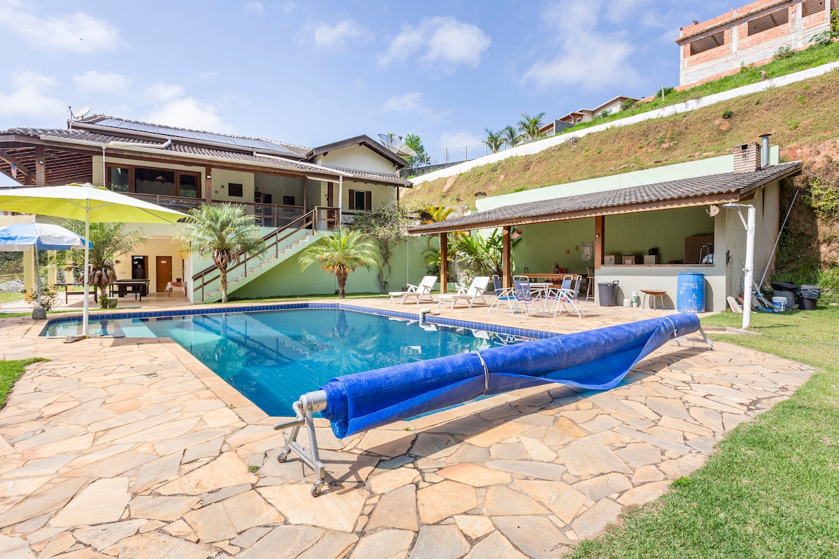 An outdoor area features a large, inviting pool surrounded by a stone patio. Sun loungers are arranged along the water's edge, shaded by large umbrellas. A covered barbecue area is visible in the background, offering space for outdoor dining and relaxation.