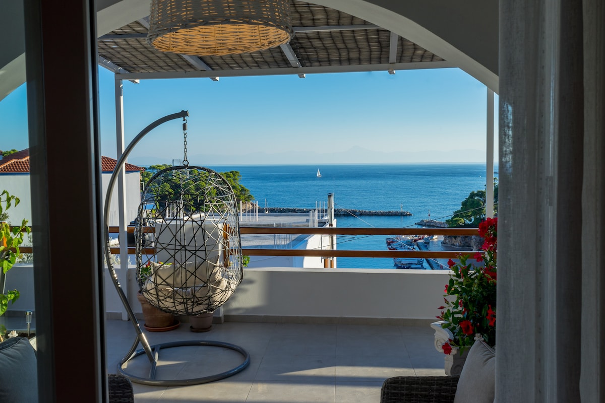 A private balcony features a hanging chair designed in an intricate spherical shape, offering a view of the sea and distant shoreline. Vibrant flowers are visible in pots, complementing the serene outdoor space bathed in natural light.