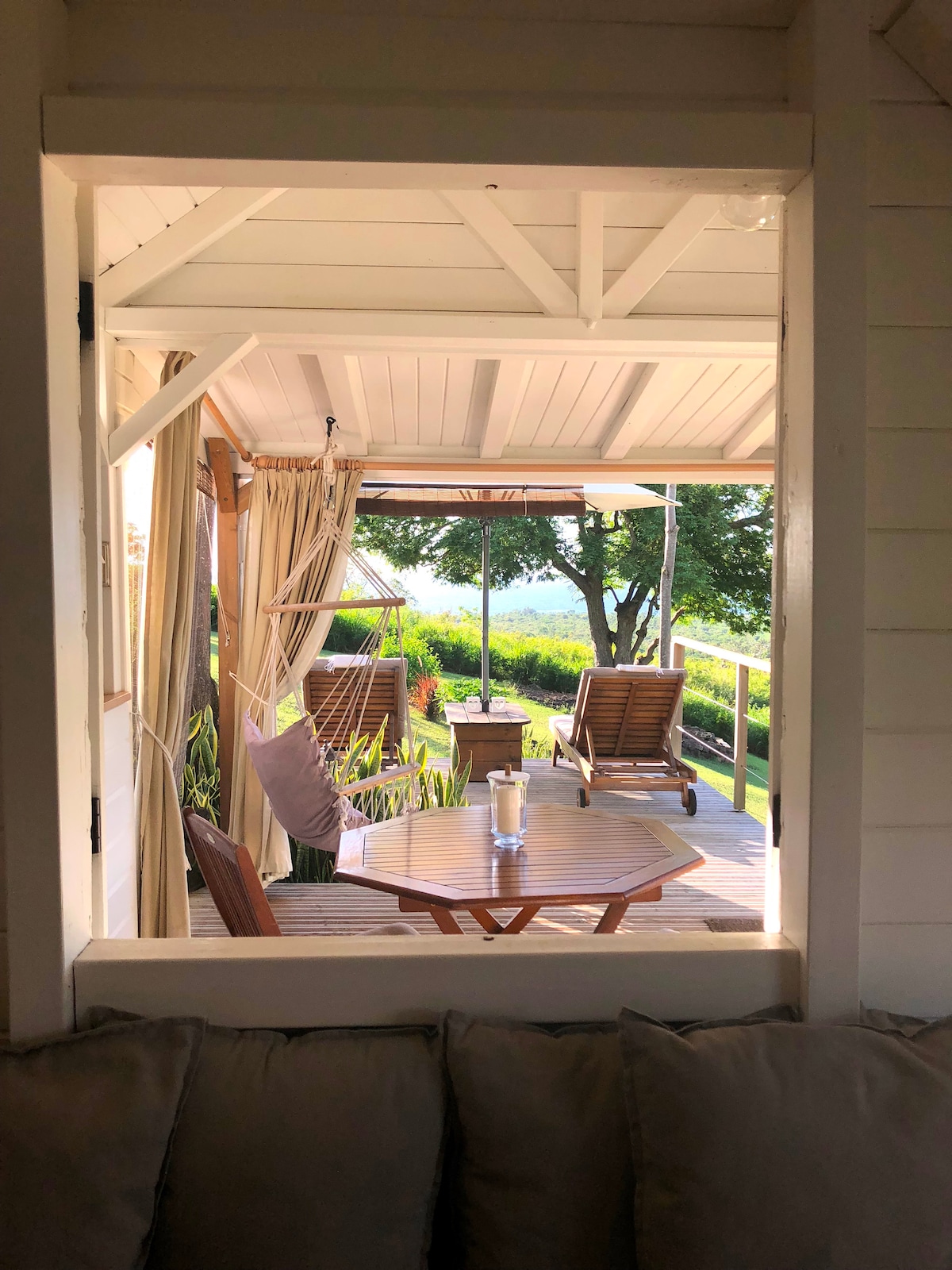 A view from an interior space frames a cozy deck area, featuring a small round table and two reclining chairs. Sunlight filters through the outdoor canopy, illuminating the surrounding greenery and a distant glimpse of the Caribbean Sea.