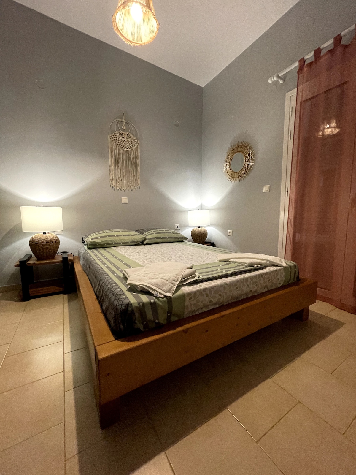 A well-lit bedroom features a spacious bed with a patterned blanket and pillows. Soft lighting is provided by bedside lamps on wooden tables. The wall is adorned with decorative wall hangings, and light filters softly through sheer curtains.