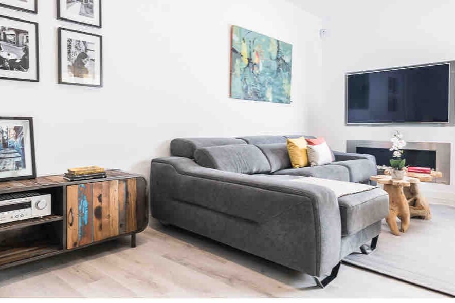 A modern living area features a grey sectional sofa adorned with colorful cushions. Artwork hangs on the white walls, complementing the natural wood side table and entertainment unit. A large flat-screen TV is mounted above a sleek entertainment center, creating a cozy yet sophisticated ambiance.