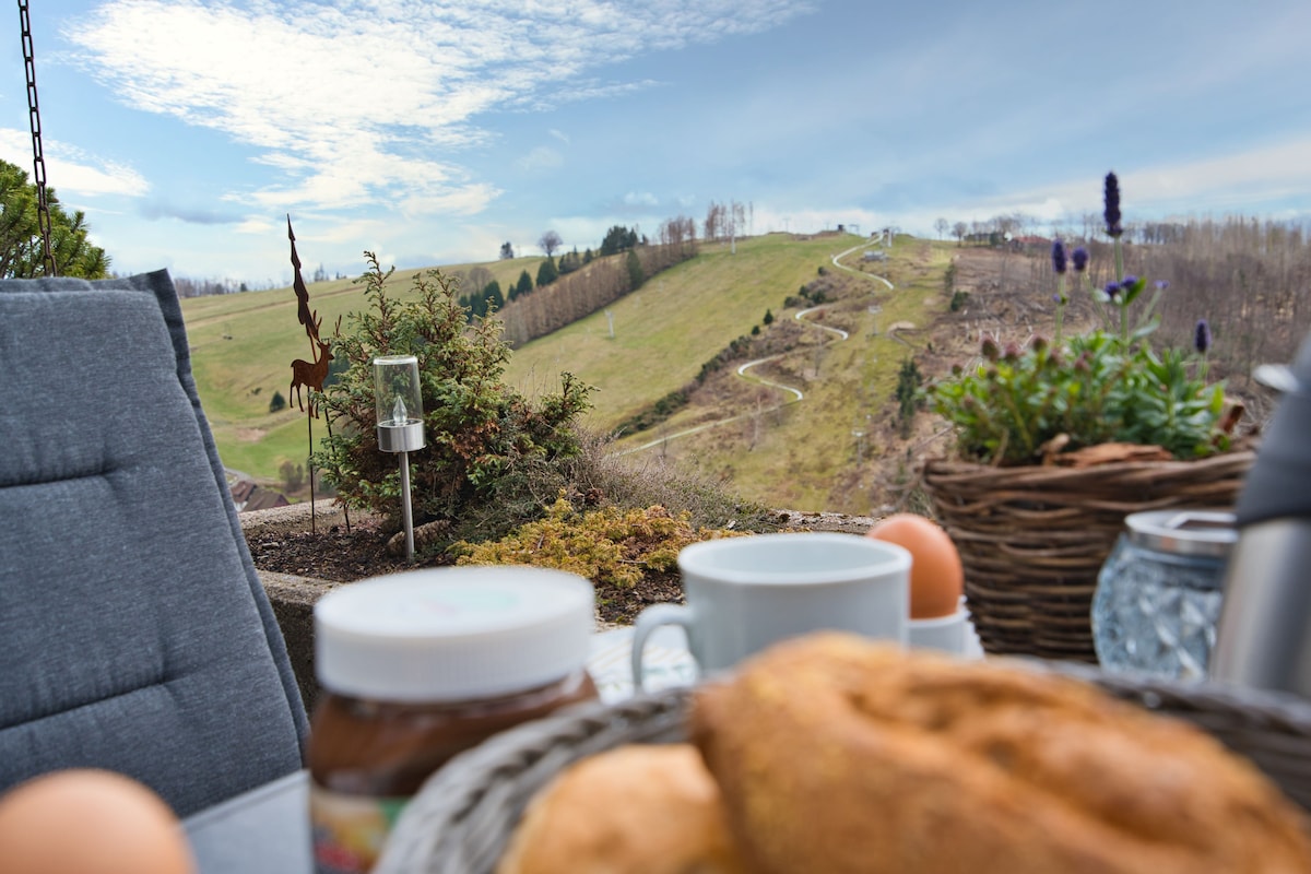 A balcony view captures rolling hills and winding paths under a cloudy sky. A cozy breakfast setting is displayed in the foreground, featuring pastries, coffee cups, and jars of spread beside decorative plants. Natural beauty and tranquility create an inviting atmosphere for leisurely mornings.