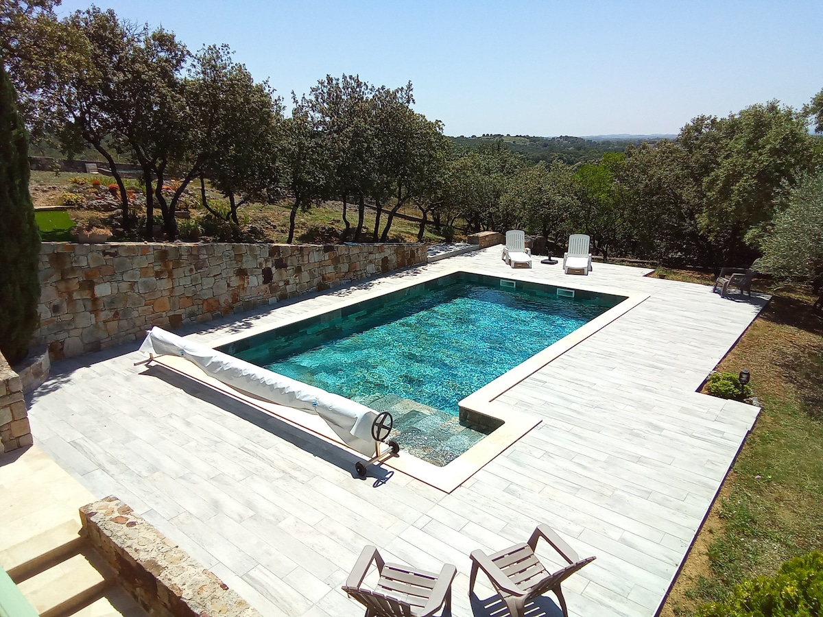 A tranquil outdoor space features a shared swimming pool surrounded by a wooden deck. Lush greenery and stone walls provide a natural backdrop. Two sun loungers are positioned nearby, inviting relaxation in a serene atmosphere.