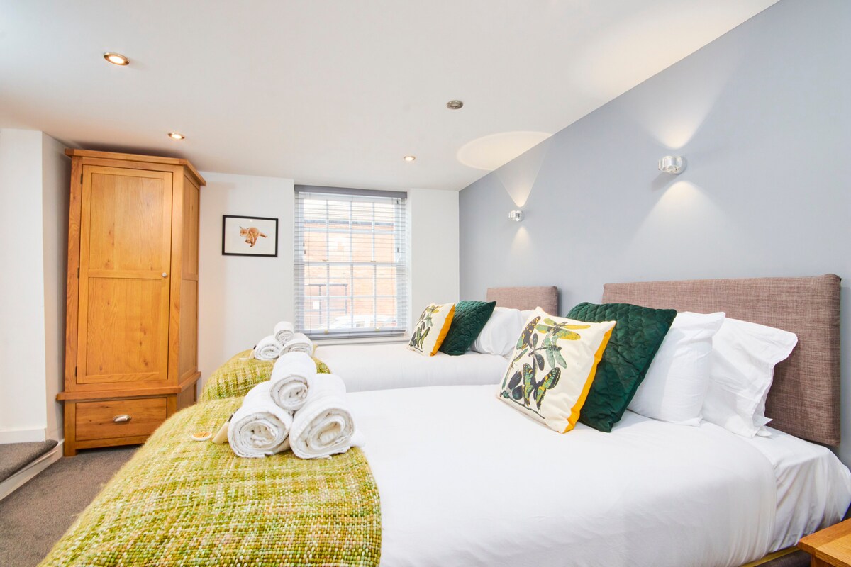 The double bedroom features two spacious beds, each adorned with decorative pillows. A vibrant green throw is draped across the beds, creating a cozy feel. Natural light enters through a window, and a wooden wardrobe sits in the corner, enhancing the room's functionality.