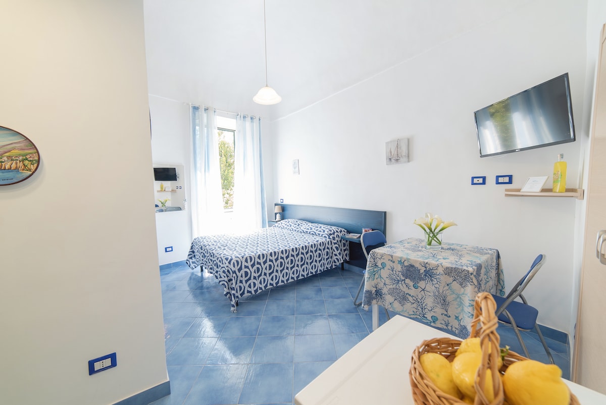 A bright and airy apartment showcases a spacious sleeping area with a patterned blue bedspread. A small dining table set for two is visible, alongside fresh fruit in a basket. Natural light filters through a window adorned with light curtains, illuminating the blue tiled floor.