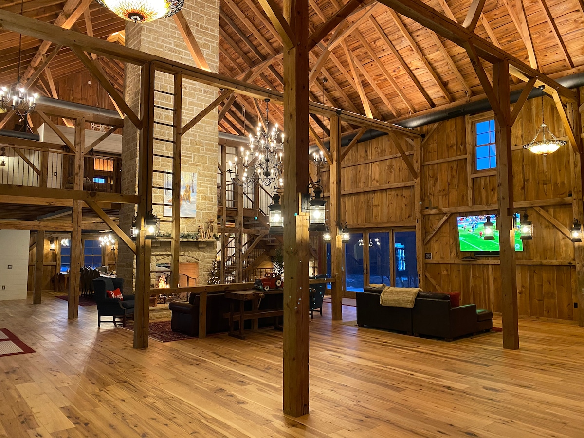 An expansive interior showcases high wooden beams and a warm, rustic ambiance. A cozy seating area is arranged around a stone fireplace, and a modern TV is mounted on the wall nearby. Large windows allow natural light to fill the open space, highlighting the wooden flooring.