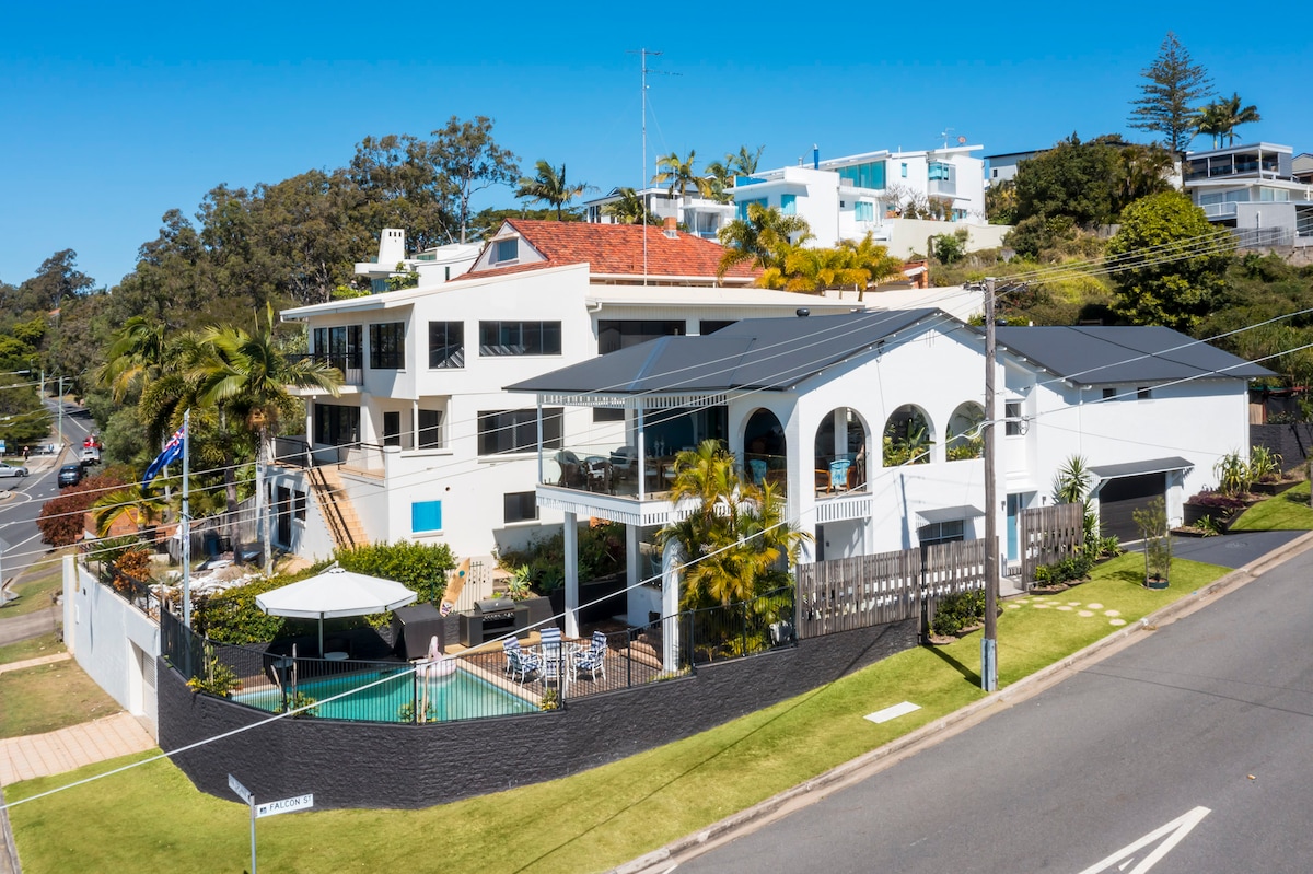 An aerial view showcases a spacious property with multiple outdoor areas, including a patio and a pool. Lush greenery and palm trees enhance the surroundings, while the architecture reflects a mix of modern and traditional styles. The nearby road provides easy access to the entrance.