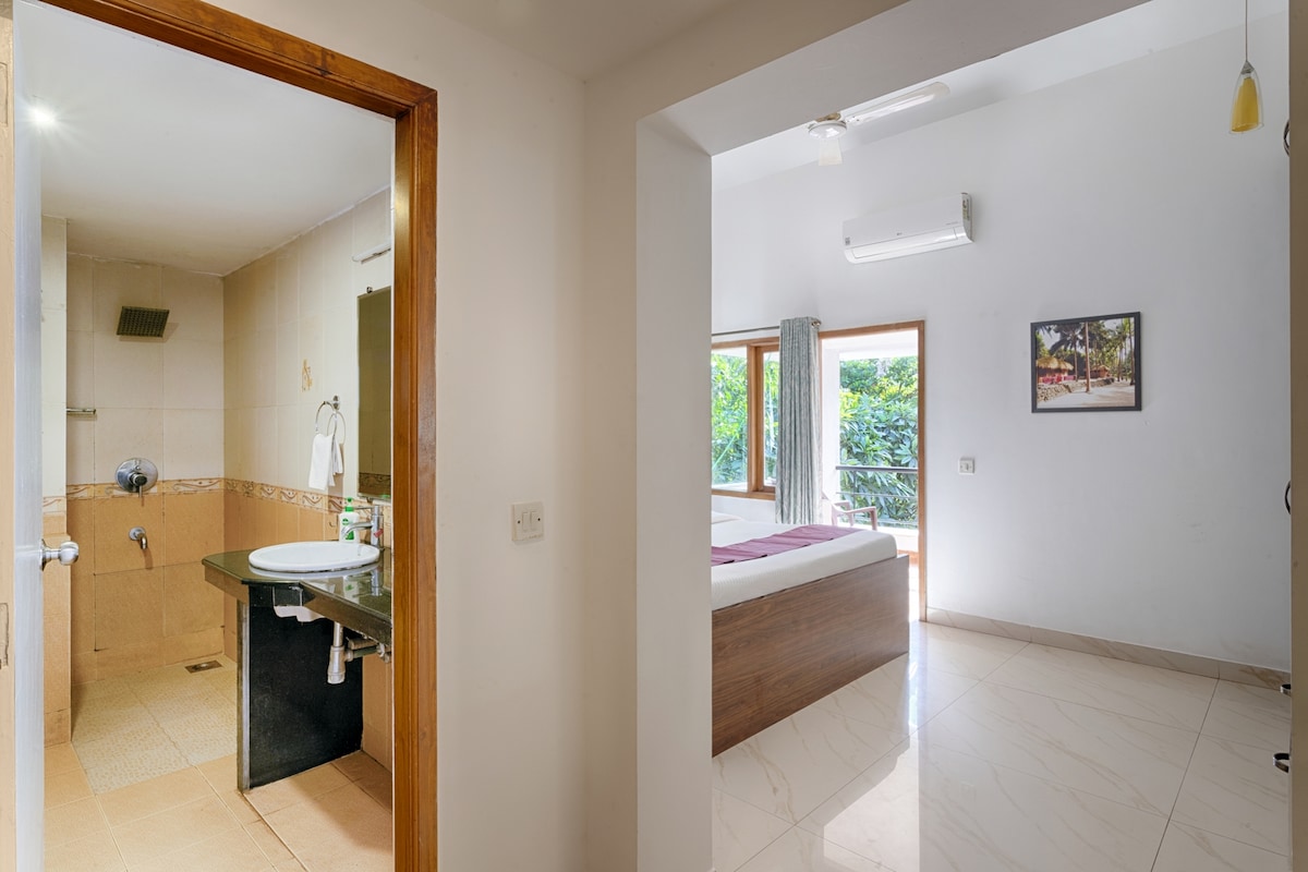 An open doorway leads from a bathroom featuring a shower and sink into a bright bedroom. The room showcases a spacious bed with light-colored linens and a large window allowing natural light to fill the space. A peaceful view of greenery is visible through the window.