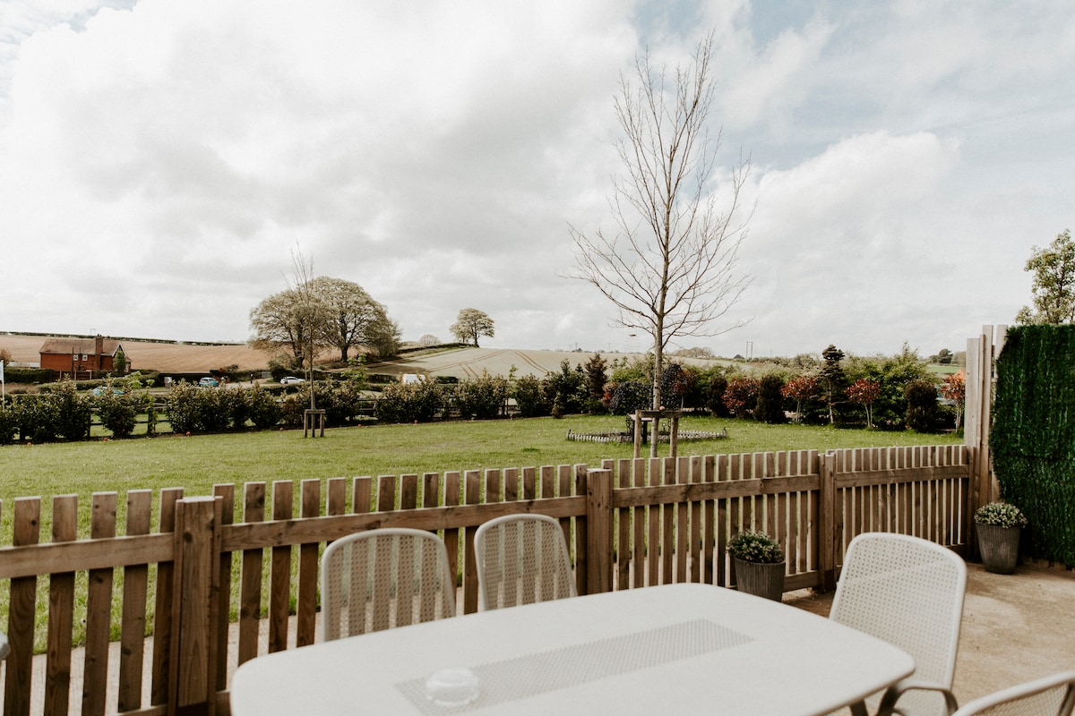 A fenced patio area features a simple outdoor table and chairs, providing a clear view of the expansive green lawn and surrounding landscape. The sky is partially cloudy, and vibrant greenery can be seen beyond the fenced area, enhancing the rural setting.
