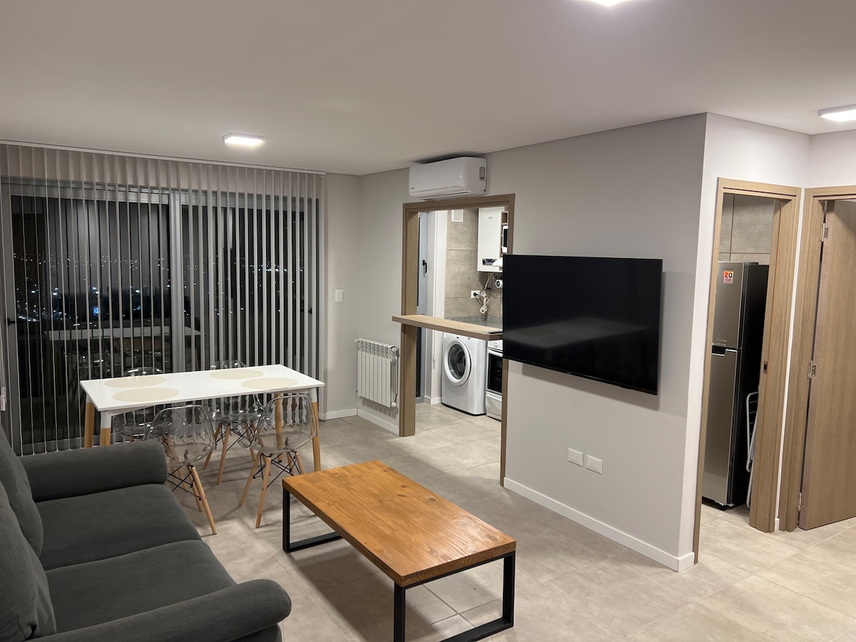 A modern living area features a cozy sofa and a wooden coffee table positioned between a dining table for four and a wall-mounted TV. A washing machine is visible in the adjacent kitchen area. Vertical blinds allow a view of the city lights through large windows.