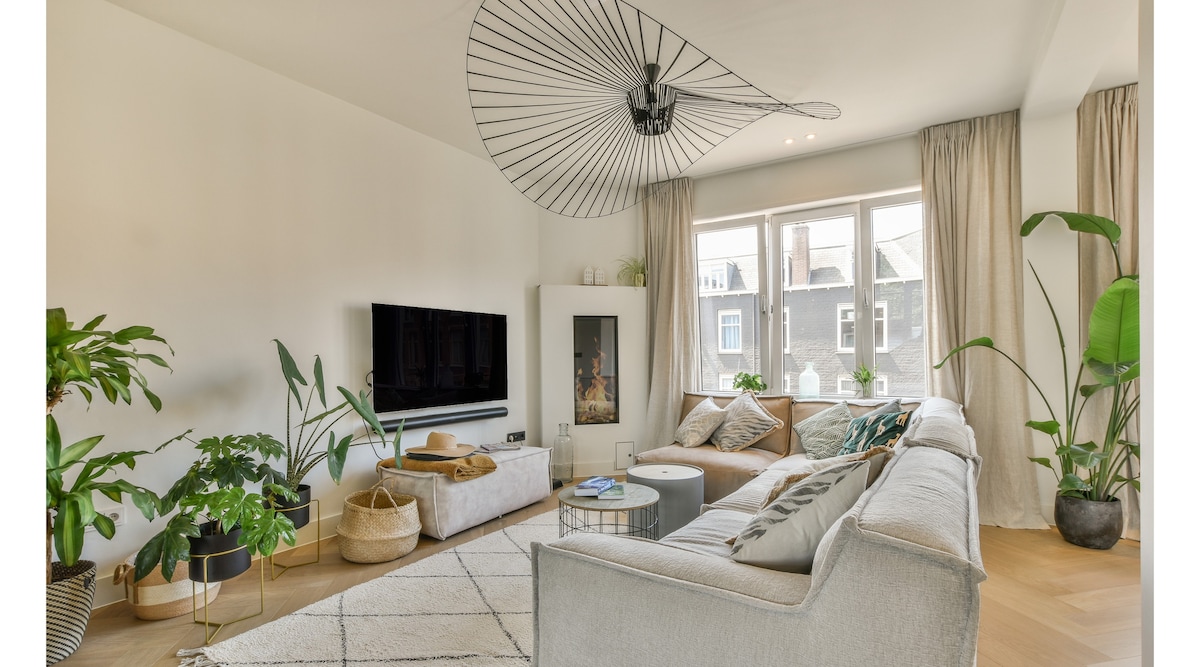 A light-filled living area features a large, L-shaped gray couch surrounded by various indoor plants. A wall-mounted television is positioned opposite, and large windows allow natural light to stream in. Soft textiles and a patterned rug add texture to the space.