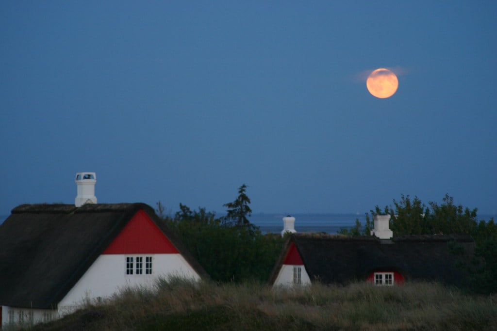 A serene scene captures two traditional thatched houses nestled among gentle dunes. A large, luminous moon hangs low in the sky, casting a soft glow over the landscape. The houses are adorned with red accents, blending harmoniously with the natural surroundings.