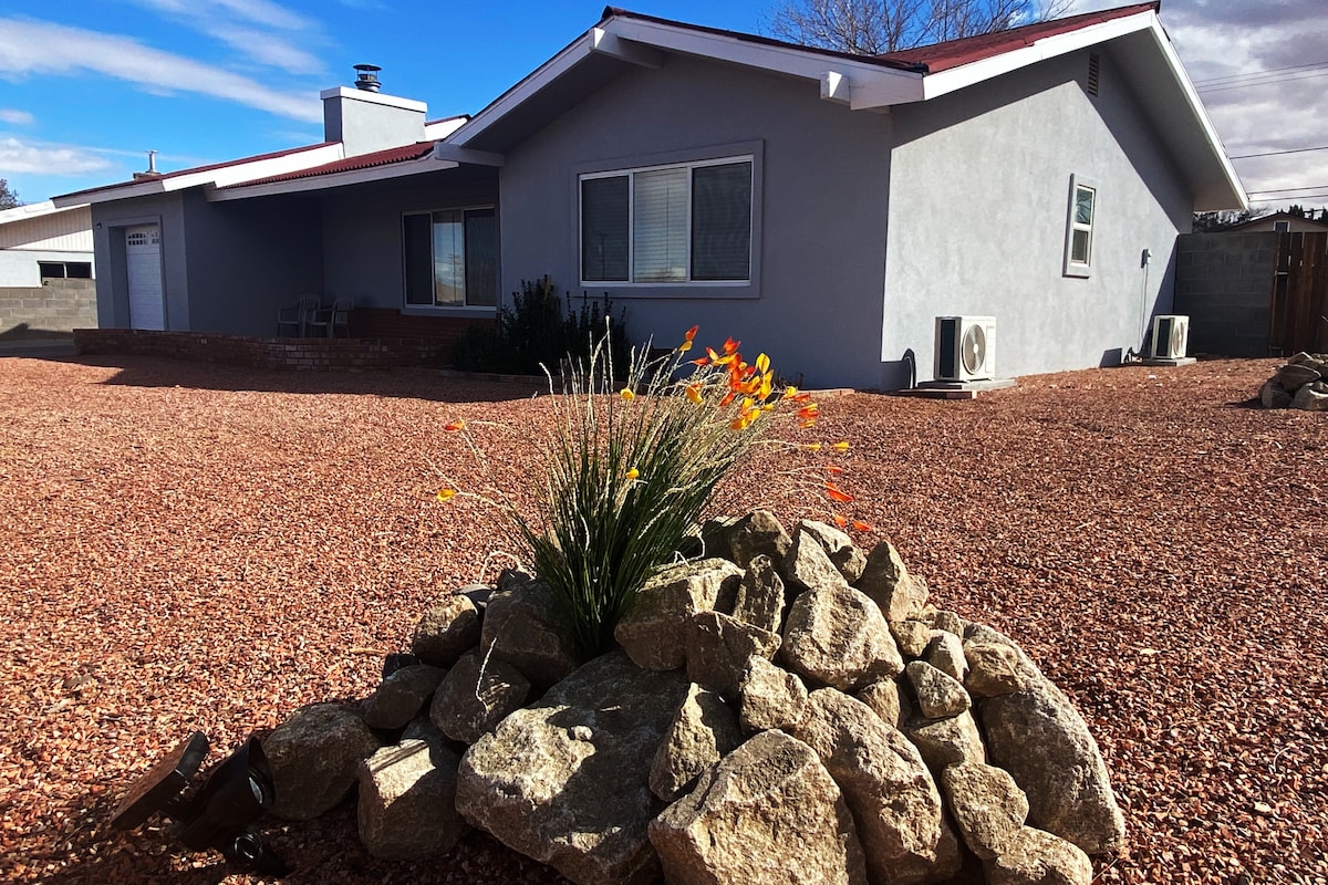 The exterior of a newly remodeled home is shown, featuring a landscaped front yard with a cluster of rocks and vibrant orange flowers. The smooth gray façade is complemented by large windows and a simple roofline, set against a gravel driveway.