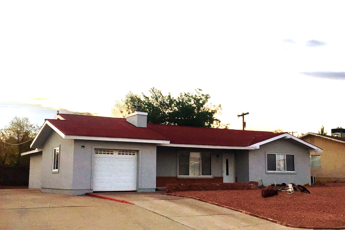The exterior of the house is showcased, featuring a modern design with a red roof and light gray siding. A garage door is visible on the left, and large windows invite natural light. The surrounding area includes a gravel driveway and sparse landscaping.