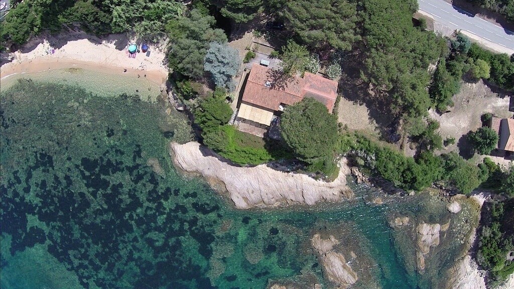 An aerial view of a coastal property nestled among trees, featuring a sandy beach nearby and rocky shoreline. The clear water displays various shades of blue and green, inviting exploration. The structure appears comfortably integrated into its natural surroundings.