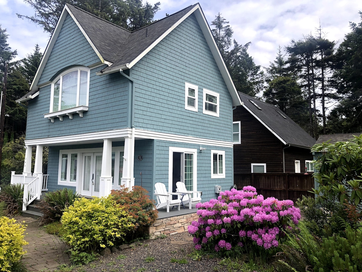 The exterior of the cottage is shown, featuring a blue wooden façade and a welcoming front porch with white chairs. Vibrant pink azaleas and lush greenery flank the stone pathway leading to the entrance, enhancing the peaceful setting among the trees.
