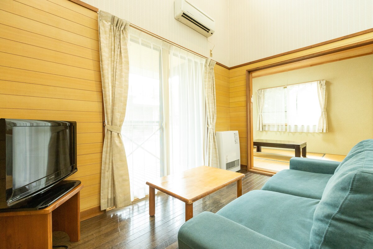 A living area features a comfortable blue sofa facing a wooden coffee table. A television is placed on a side cabinet, while sheer curtains frame a window allowing natural light to fill the space. A separate area with a reflective surface is visible in the background.