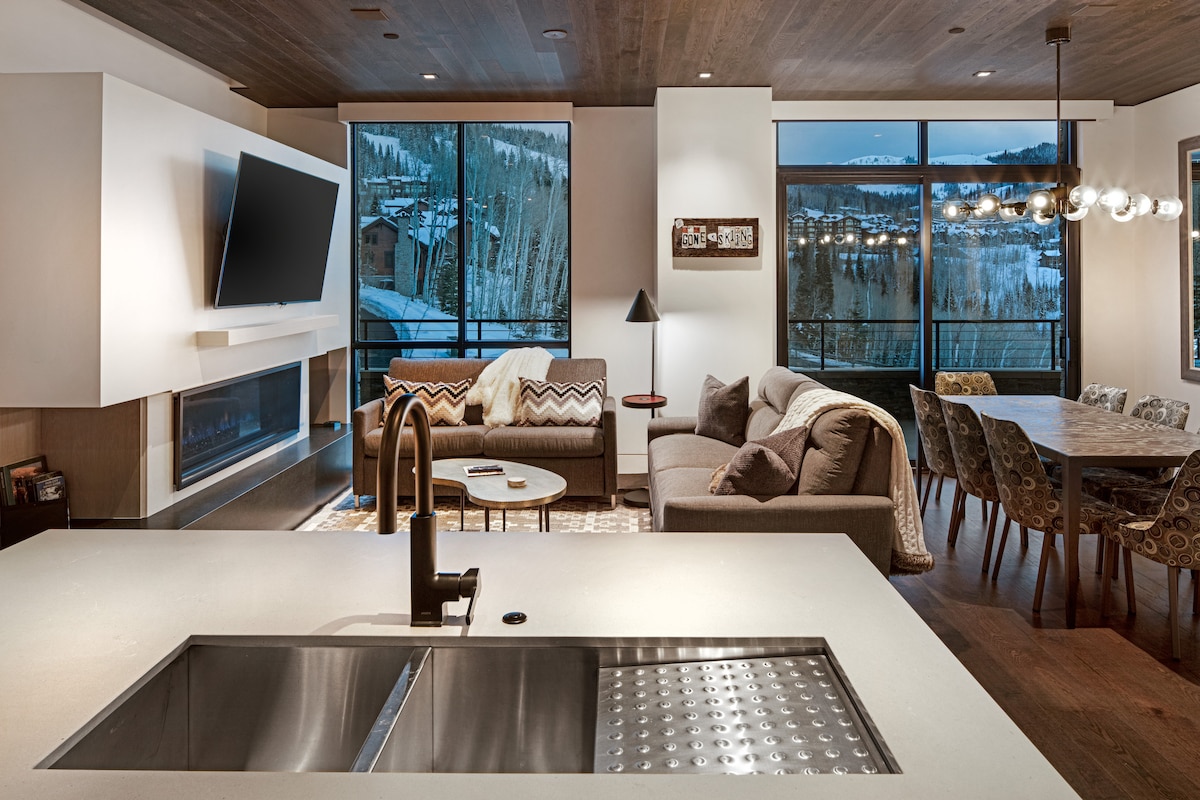 A modern living area showcases a sectional sofa and decorative armchair against large windows revealing mountain views. A sleek kitchen island features a stainless steel sink. A wooden dining table with patterned chairs is positioned nearby, while a soft rug adds warmth to the space.