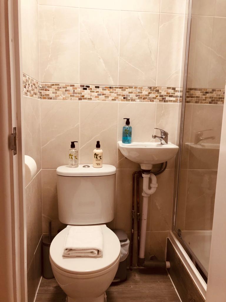 A modern shower room is equipped with a toilet, a small sink, and a shower enclosure. Various toiletries are neatly arranged on the sink, and a folded towel is placed on the toilet's tank. Neutral tiles create a clean and contemporary ambiance.
