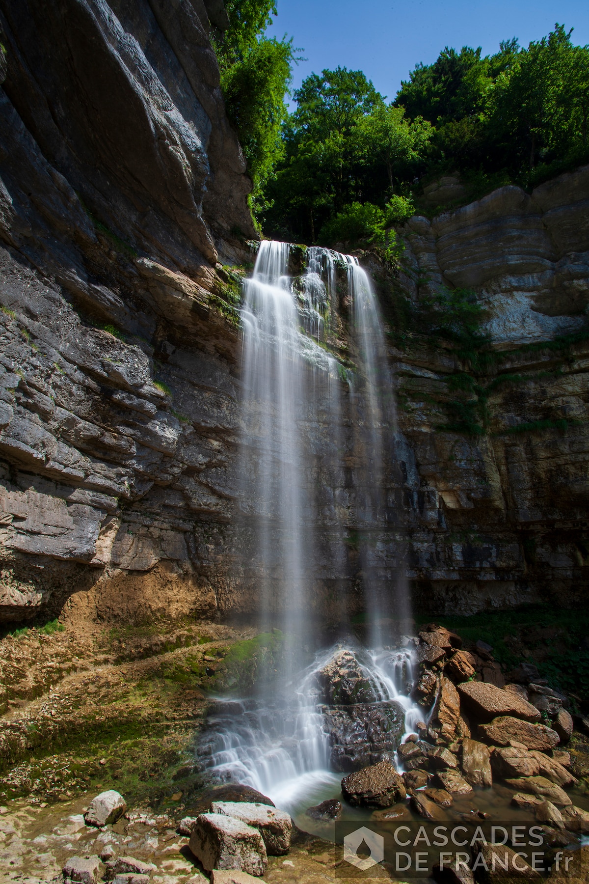 A waterfall cascades down a rocky cliffside, creating a mist at its base. Surrounding lush greenery enhances the natural setting, while large boulders rest in the foreground. The sunlight reflects on the water, highlighting the textures of the stone and the flow of the waterfall.