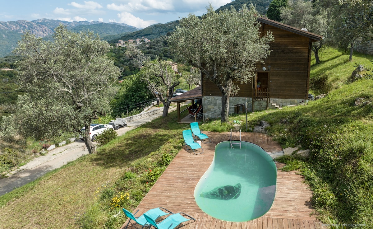 A private pool is positioned on a wooden deck, surrounded by greenery and olive trees. A rustic chalet is visible in the background, nestled against a mountainous landscape. Lounge chairs in soft blue complement the natural setting, inviting relaxation.