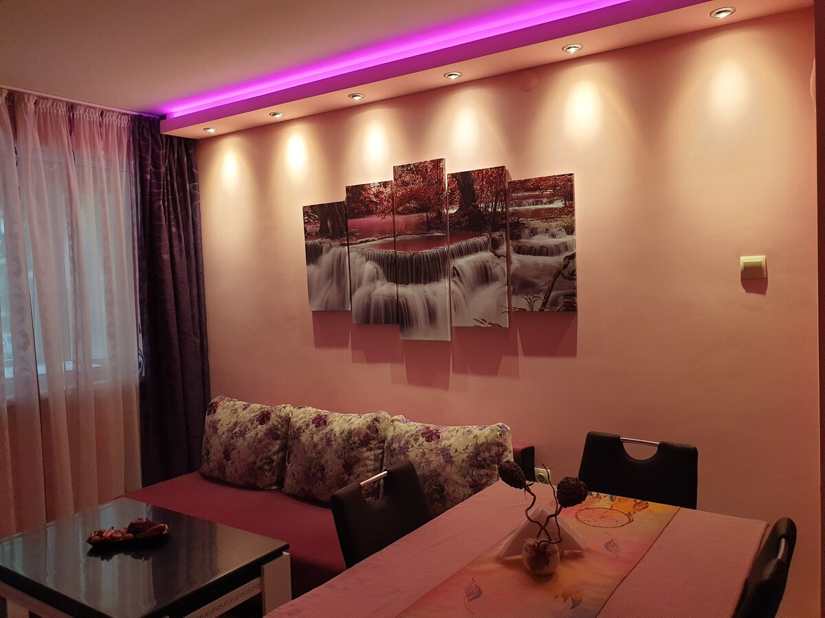 A comfortable living area is highlighted with soft pink walls and ambient LED lighting. A decorative canvas featuring a serene water scene is displayed above a cushioned seating area. A dining table is positioned nearby, complemented by simple decor and floral accents.