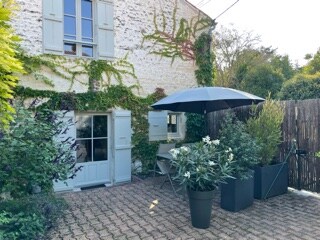 An outdoor area features a cozy seating arrangement shaded by a large umbrella, surrounded by potted plants and vibrant greenery. The quaint façade of the building is adorned with climbing vines, enhancing the tranquil atmosphere of the private courtyard.