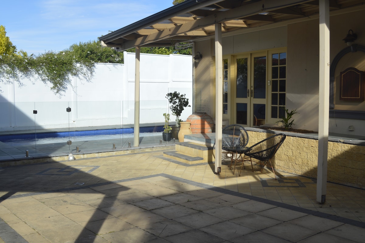 A patio area is visible, featuring a stone floor arrangement and two wicker chairs positioned nearby. A swimming pool is shown in the background, enclosed by a glass barrier. The space is bathed in natural light with greenery visible beyond the pool area.