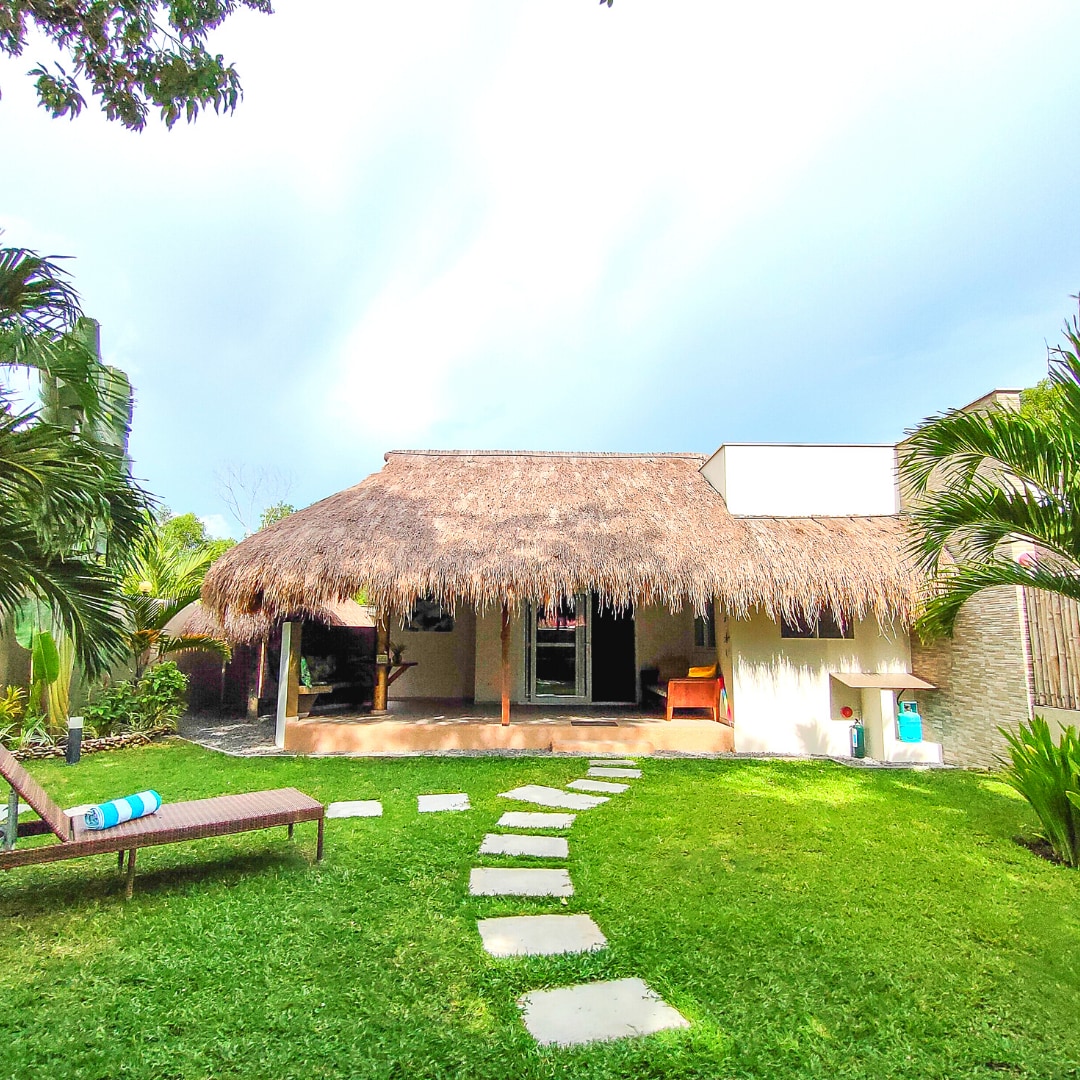 A private villa is nestled among lush tropical gardens, featuring a thatched roof and a covered terrace. A pathway of stepping stones leads to the entrance, while sun loungers are positioned on the well-kept lawn, offering a serene outdoor experience.