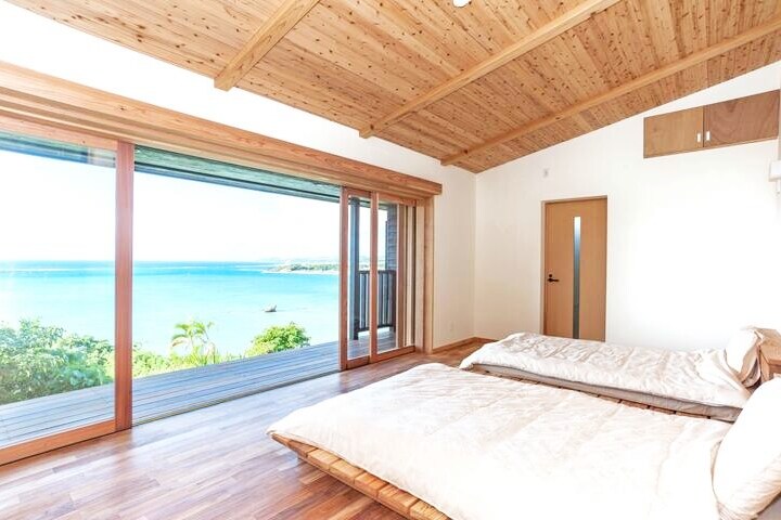 A bright room features two spacious beds positioned on a wood floor, with large sliding glass doors providing panoramic views of the sea and surrounding landscape. Natural light fills the space, and wooden beams are visible on the ceiling, enhancing the airy atmosphere.