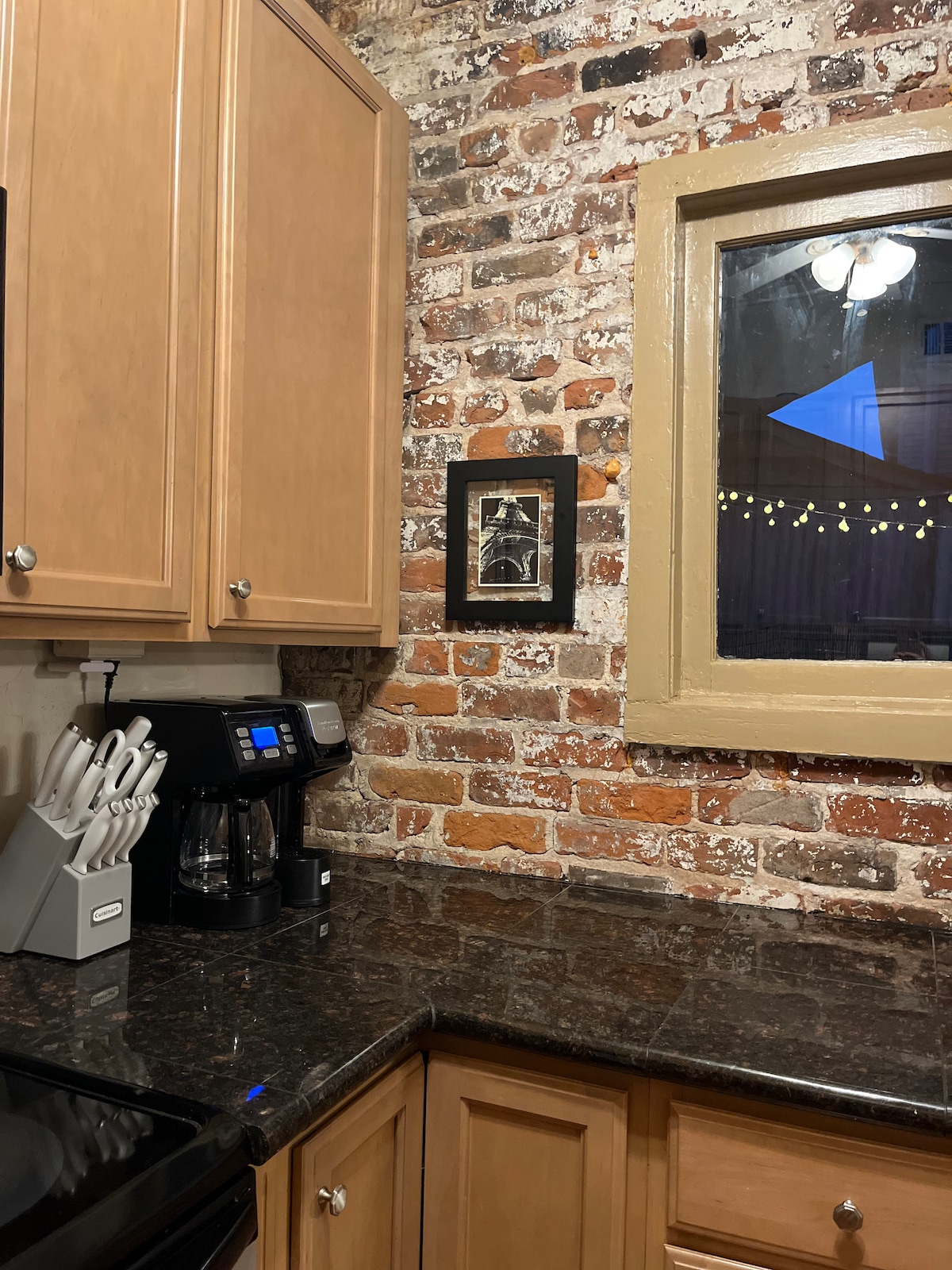 A kitchen corner features exposed brick walls, creating a rustic ambiance. Wooden cabinets house essential items, while a dark countertop contrasts with the warm wood tones. A coffee maker is positioned nearby, and a window allows natural light to illuminate the space.