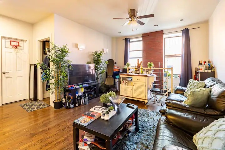 A spacious living area features a comfortable black leather sofa and a coffee table adorned with various items. A desk and chair are positioned near a large window, while a television stands on a console. Plants add greenery, enhancing the atmosphere of the room.