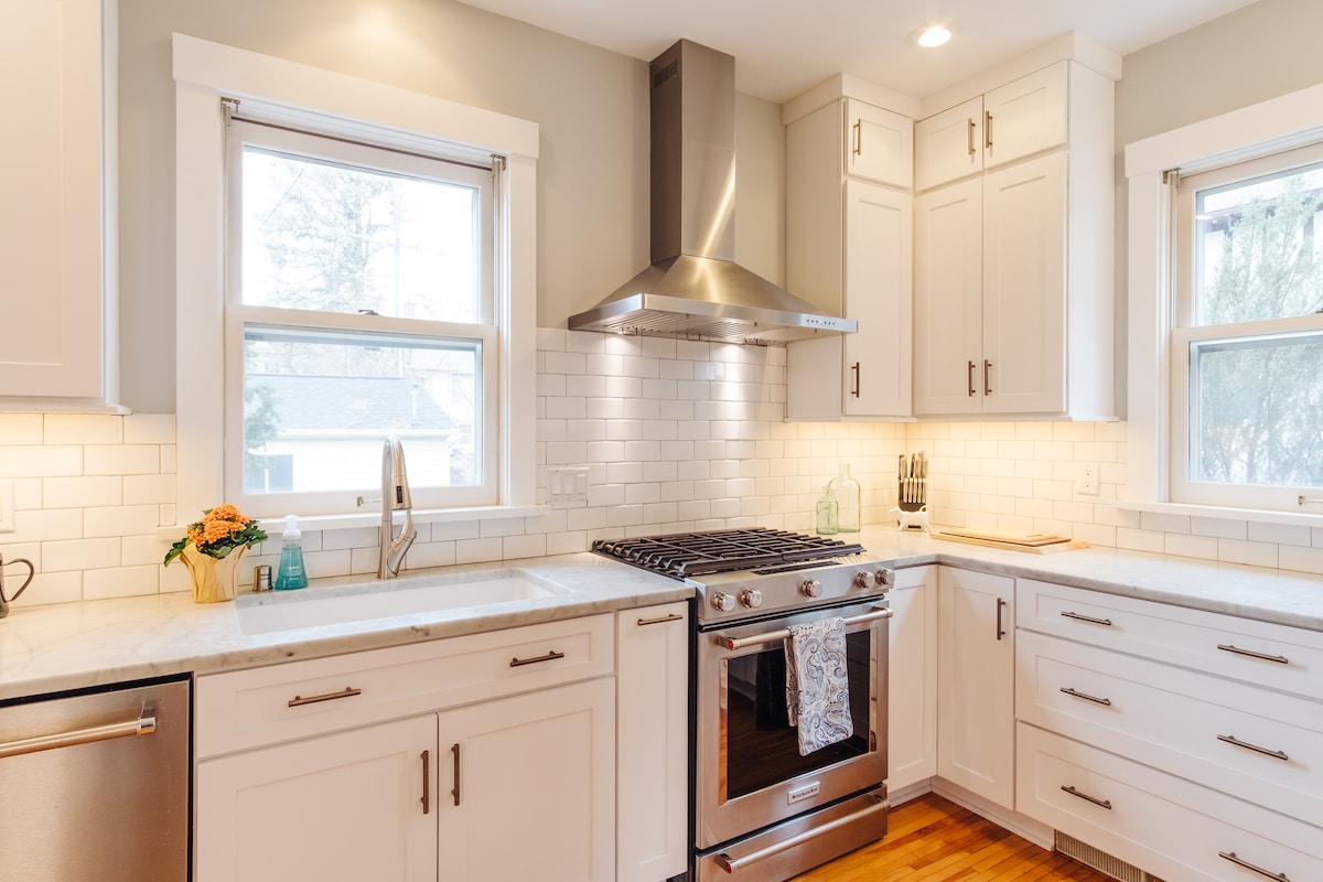A modern kitchen is featured, equipped with high-end stainless steel appliances and elegant cabinetry. Marble countertops are complemented by a stylish backsplash. Natural light streams in through two windows, illuminating a spacious sink and additional countertop space, enhancing functionality.