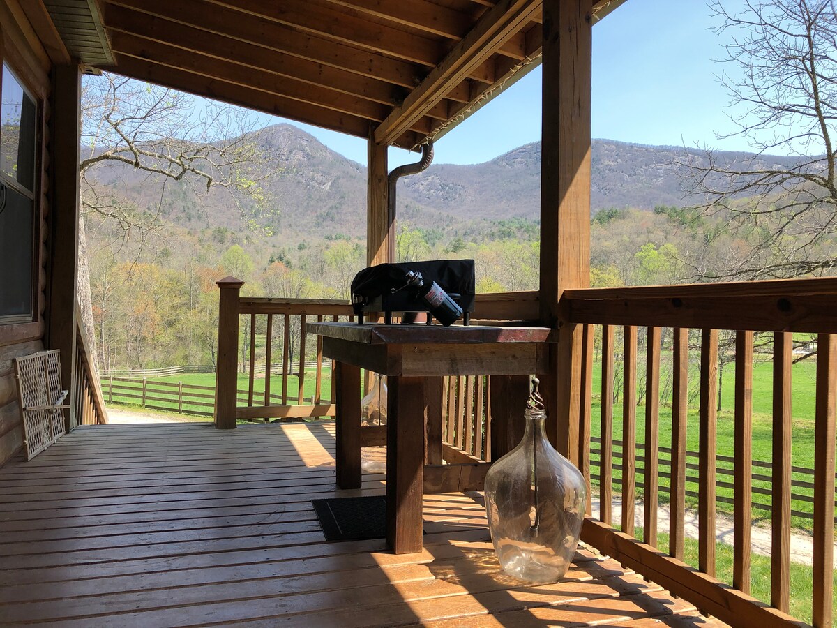 The front porch features a sturdy wooden floor and a rustic table, with views of the surrounding mountains in the background. A decorative glass jar rests on the floor, enhancing the outdoor space's natural charm while overlooking a vibrant lawn.