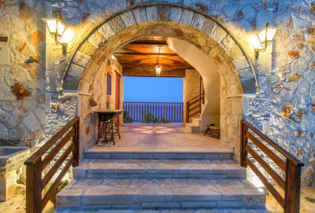 The entrance features a stone archway, with warm lights illuminating the area. Steps lead to a spacious landing that offers views of the surrounding landscape. A rustic wooden table is positioned against the stone wall, creating a welcoming transition to the interior.