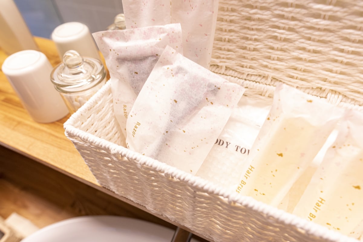 A neatly arranged white woven basket holds individually wrapped toiletries, including body towels and other personal care items. The wooden surface beneath adds a natural touch, complementing the overall cleanliness and organization of the space.