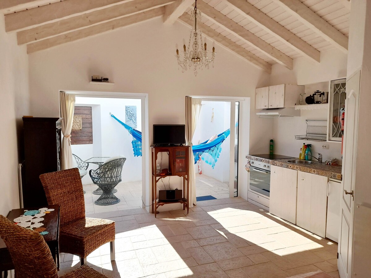A bright and airy interior features an open layout with a kitchenette on one side. Comfortable seating is arranged around a small table, complemented by a TV. Large glass doors lead to a private outdoor space with hammocks, inviting natural light into the room.