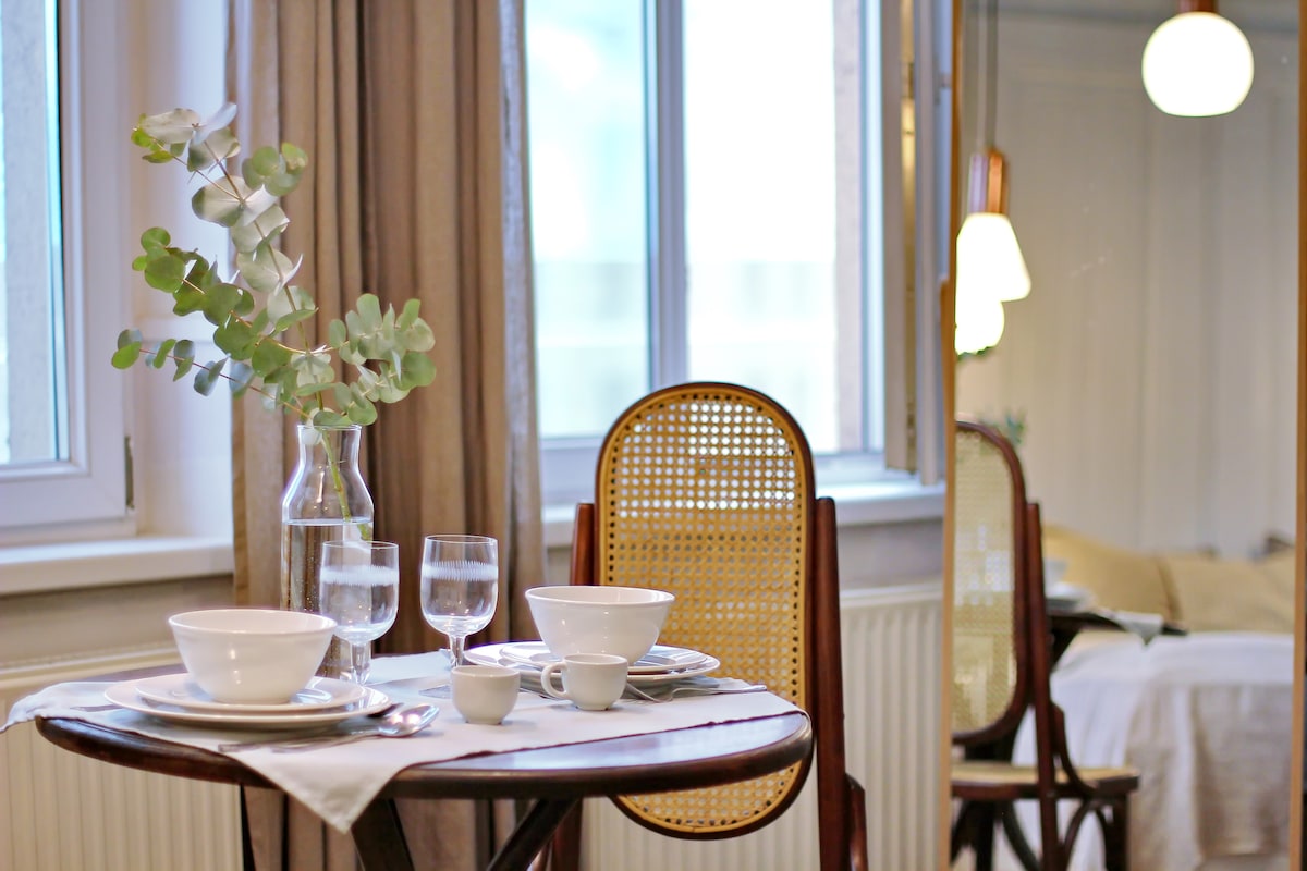 A dining table is set for two, featuring white dishware and glassware beside a vase with fresh greenery. Natural light filters through nearby windows, and the reflection shows a cozy sleeping area with soft bedding.