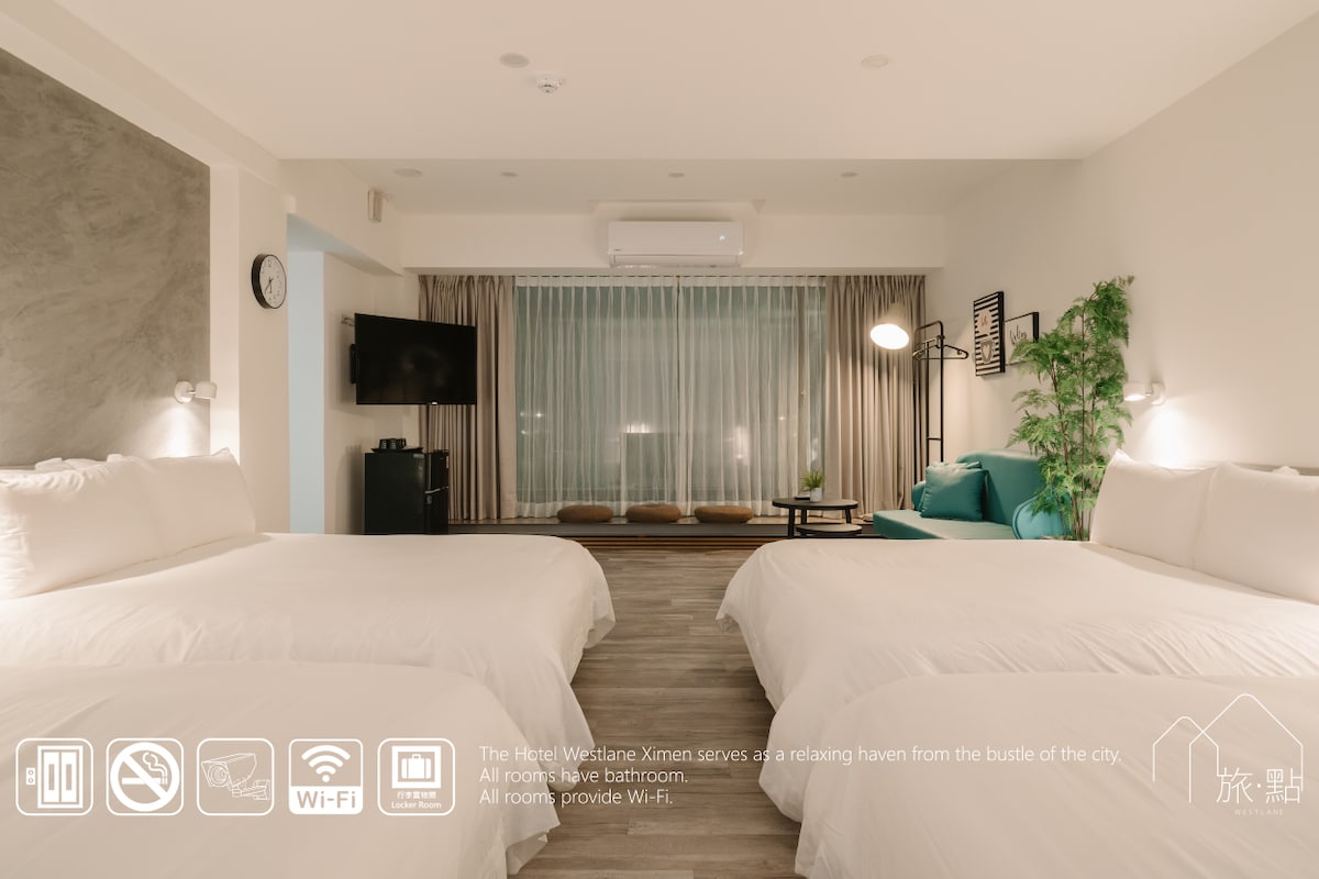 A bright suite features two spacious beds with crisp white linens. A large glass window allows natural light to fill the room, complemented by sheer curtains. A cozy seating area with a green couch and a wall-mounted television is visible, creating a welcoming environment.