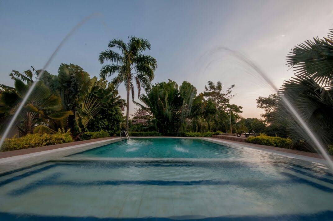 A serene pool area is surrounded by lush greenery, featuring aquatic fountains that create a gentle spray. Towering palm trees and tropical plants frame the scene, enhancing the natural beauty of the space as the sun sets in the background.