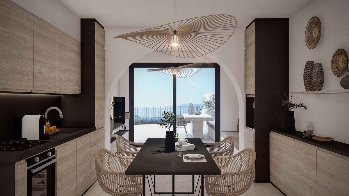 The kitchen features modern cabinetry with a light wood finish and dark accents. A spacious dining table is surrounded by woven chairs, providing a relaxed atmosphere. Large glass doors ensure ample natural light and offer views of the terrace and sea beyond.