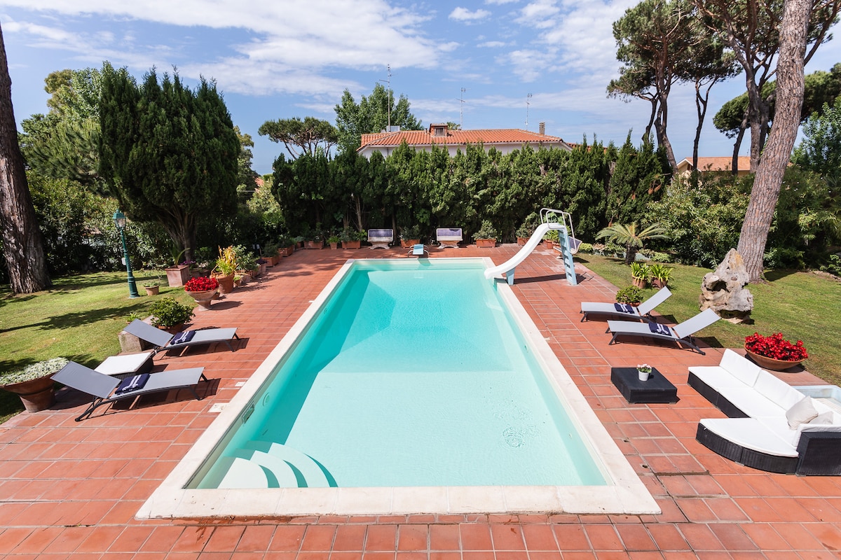 An inviting pool area features a clear swimming pool equipped with a slide, surrounded by landscaped gardens and tall trees. Lounge chairs are arranged around the pool, offering a tranquil space for relaxation. A shaded area is positioned nearby for comfortable gatherings.