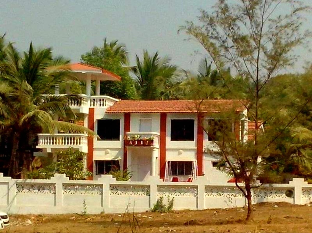 A two-story villa is framed by lush palm trees, showcasing a terracotta-tiled roof and white walls. A balcony on the upper level offers outdoor seating, while the garden area features green foliage. An ornamental fence surrounds the property, enhancing its spacious setting.