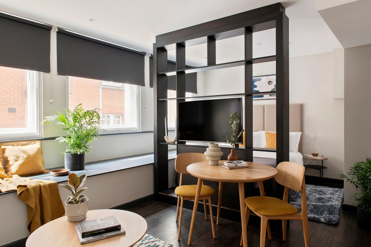 A stylish studio apartment features an open layout with a wooden dining table and two yellow chairs. Shelving units frame a large flat-screen TV, while large windows allow natural light to brighten the space. A cozy bed is visible in the background, complemented by decorative cushions.
