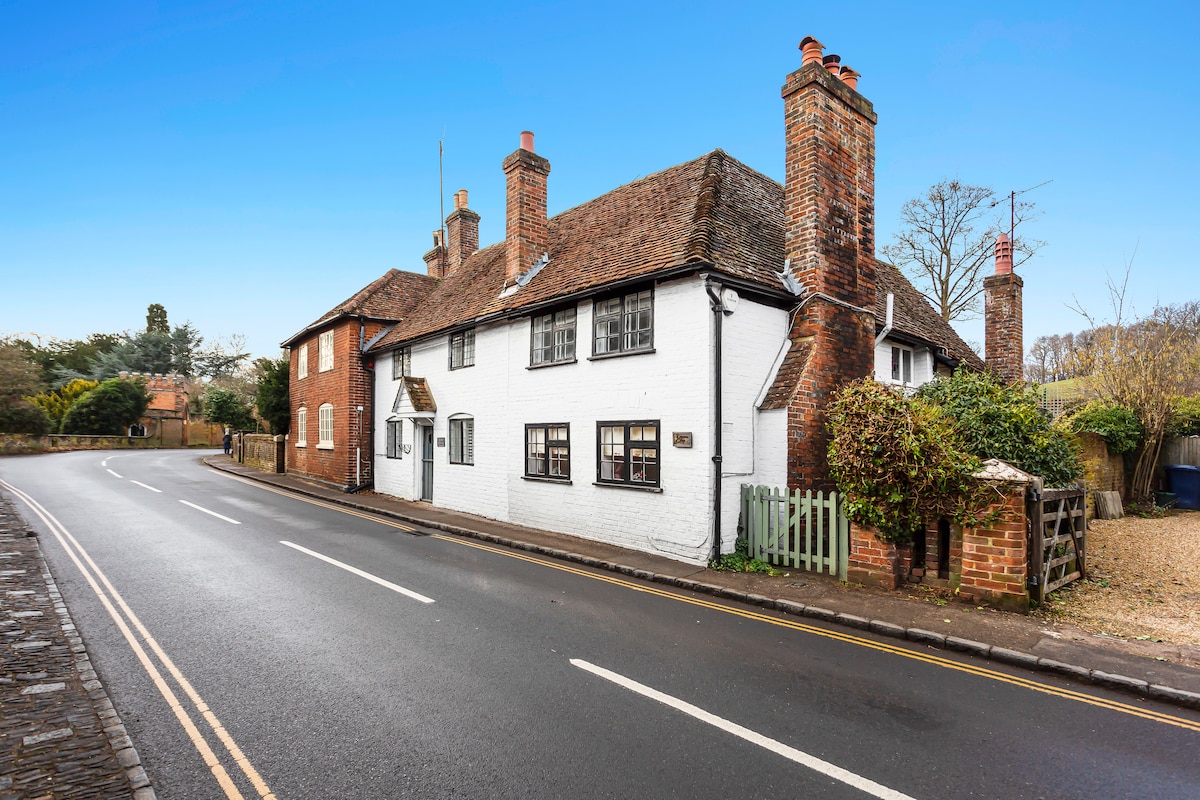 A charming timber-framed cottage is viewed from the street, featuring a white exterior and multiple chimneys. The property is framed by greenery, with a green gate marking the entrance. The peaceful surroundings reflect the tranquil nature of the village.
