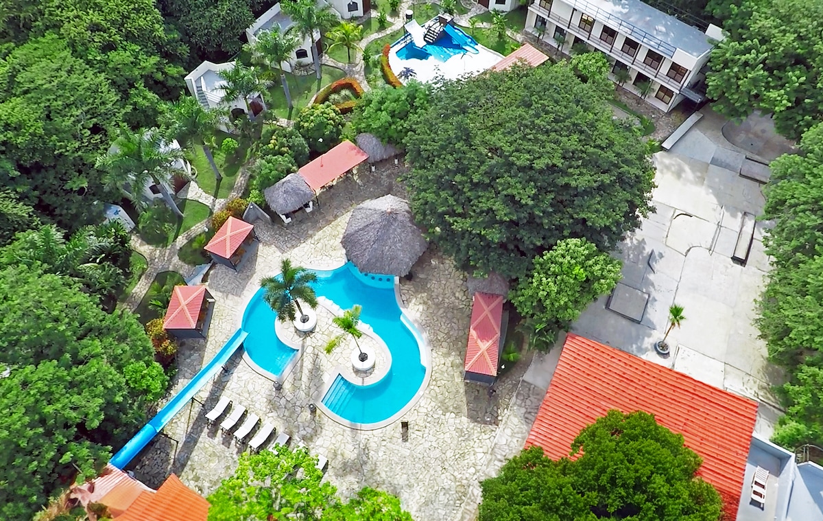 Aerial view of the Surf Ranch Resort showcases the expansive pool area, surrounded by lush greenery and palm trees. A waterslide blends into the pool, bordered by lounge chairs. The resort buildings are visible in the background, complemented by vibrant red roofs.