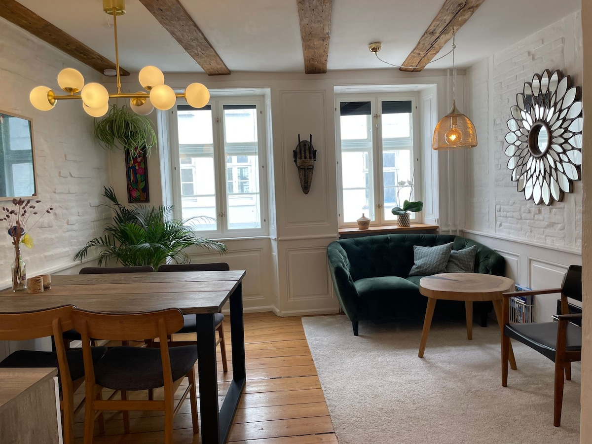 A cozy living space features a green sofa paired with a circular wooden table and chairs. Natural light streams through two windows dressed with simple curtains. Decorative plants and a mirror add visual interest to the bright, inviting interior, highlighting the wooden beams on the ceiling.