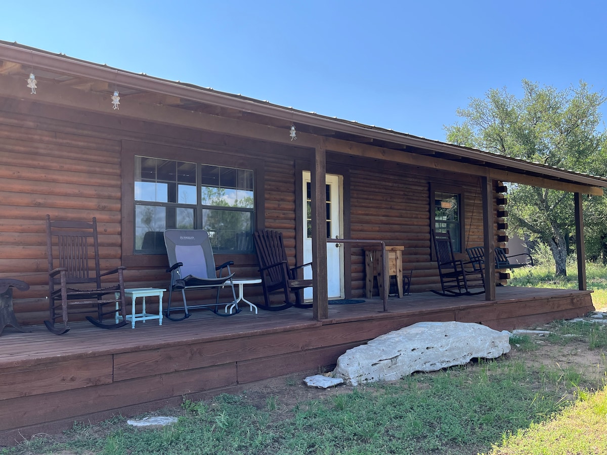 A rustic cabin porch showcases two rocking chairs and a small table, inviting moments of relaxation. A smooth stone rests on the ground, while greenery surrounds the area. Large windows provide views of the outdoors, enhancing the cabin's connection to nature.